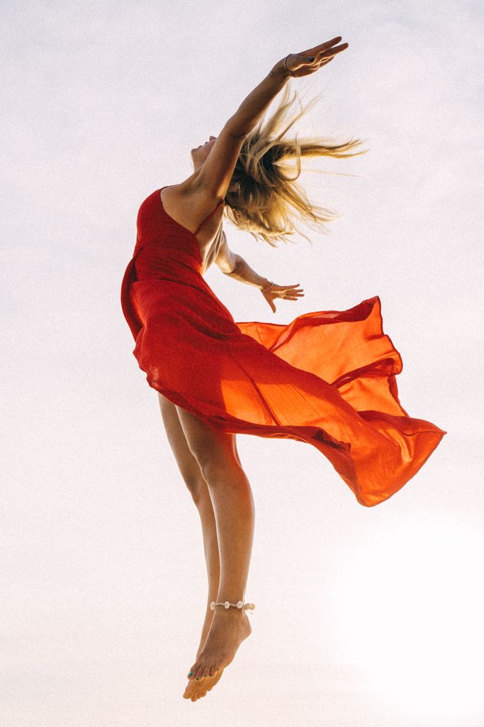 A woman in a flowing red dress elegantly jumps with arms raised against a bright sky.